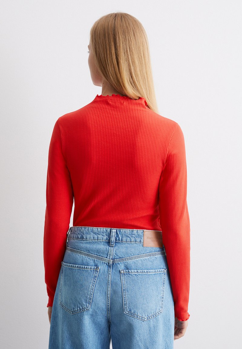 Rotes geripptes Langarmoberteil mit einem gewellten Ausschnitt, kombiniert mit hochgeschnittenen hellblauen Jeans aus Denim mit Gesäßtaschen und einem Lederetikett.