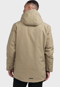 Beige wasserdichte Jacke mit hohem Kragen und großer Kapuze, flachem Rückenbund und dezentem Branding am unteren Rücken.