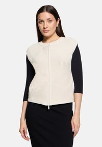 Veste sans manches en maille crème avec fermeture éclair à l'avant, présentant des motifs texturés et un col rond, portée sur un haut noir à manches longues.