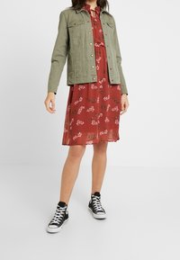 Chaqueta de mezclilla verde sobre un vestido rojo con flores y mangas cortas, combinado con zapatillas negras altas. El vestido tiene un escote con volantes.