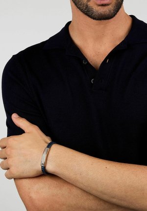 Man met een zwart poloshirt met korte mouwen en een blauwe gevlochten armband met een rechthoekige metalen sluiting om zijn pols.