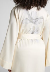 Vrouw draagt een crèmekleurige satijnen robe met brede mouwen en een groot subtiel bloemmotief op de achterkant, vastgebonden in de taille met een bijpassende riem.