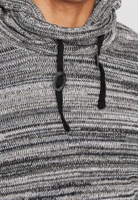 Sudadera de punto a rayas grises y negras con capucha, cuello vuelto y cordones. Textura suave con fibras contrastantes por todo el tejido. Destacan los acentos negros.