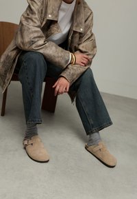 Beige suède instap schoenen met een plat voetbed en een zijgesp, gecombineerd met grijze sokken en denim jeans. Het model zit op een houten stoel.