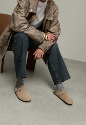 Beige suède instap schoenen met een plat voetbed en een zijgesp, gecombineerd met grijze sokken en denim jeans. Het model zit op een houten stoel.