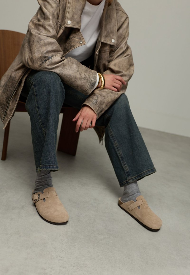 Beige suède instap schoenen met een plat voetbed en een zijgesp, gecombineerd met grijze sokken en denim jeans. Het model zit op een houten stoel.