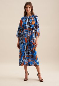 Blaues Blumenkleid mit Puffärmeln, orange und schwarzen Mustern, tailliertem Oberteil und knielanger Länge. Schwarze Absatzsandalen runden den Look ab.