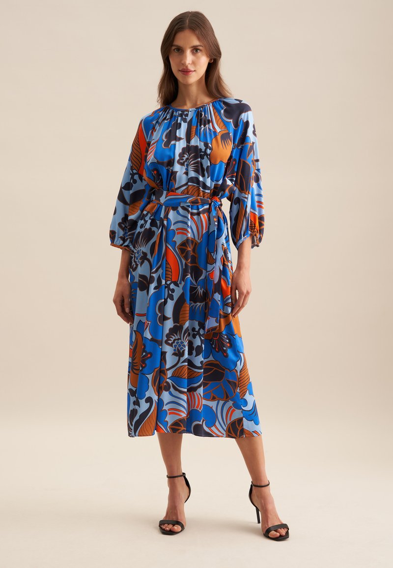 Blaues Blumenkleid mit Puffärmeln, orange und schwarzen Mustern, tailliertem Oberteil und knielanger Länge. Schwarze Absatzsandalen runden den Look ab.
