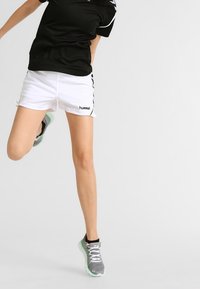 Svart och vit träningsklädsel består av en svart topp med kort ärm och vita shorts. Shortsen har en logotyp och sidosträck. Grå träningsskor är på fötterna.