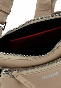 Beige leren schoudertas met zilver hardware, voorzien van een ritsvak gevoerd met zwart en een rode kaarthouder binnenin. Logo zichtbaar.