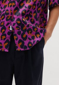 Camicetta corta viola con motivo leopardato caratterizzato da accenti neri e arancioni. Abbinata a pantaloni scuri, evidenziando i dettagli dei bottoni.