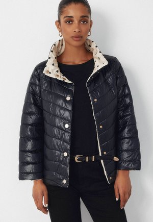 Kobieta ubrana w czarną pikowaną kurtkę z kremową podszewką w cętki, czarny top, czarne spodnie oraz czarny pasek ze złotą klamrą, stojąca na tle białej ściany.