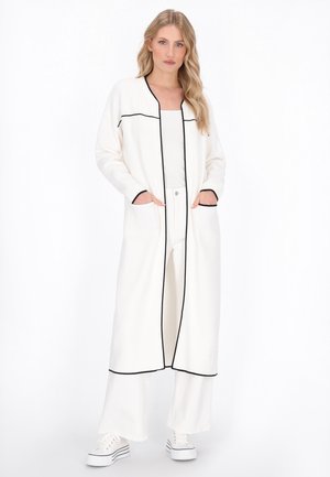 Lange witte coat met zwarte bies, open voorkant, twee zijzakken, gedragen over een lichte top en brede broeken, gecombineerd met witte sneakers.