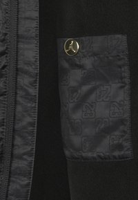Sort fleece beklædning med lynlås, der har en tekstureret lomme med et subtilt logo mønster og en guldknap detalje.