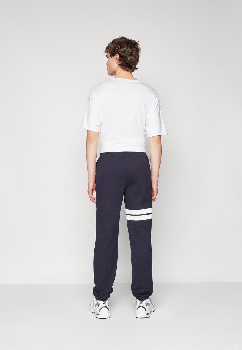 Pantalons de survêtement bleu marine avec des rayures blanches sur la jambe gauche, avec des poignets et une taille élastiques, associés à un T-shirt blanc à manches courtes.