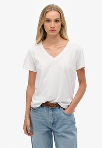 Mujer con cabello largo y rubio que lleva una camiseta blanca de cuello en V metida dentro de unos vaqueros azul claro con un cinturón trenzado marrón, de pie con una mano en el bolsillo.
