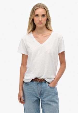 Vrouw met lang blond haar, gekleed in een witte V-hals T-shirt, ingestopt in lichtblauwe jeans met een bruine gevlochten riem, staande met één hand in de zak.