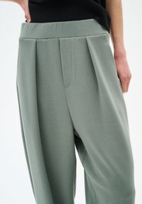 Pantaloni larghi di colore verde chiaro, con pinces, caratterizzati da una texture liscia e un alto punto vita. Tasca laterale visibile sul lato sinistro.