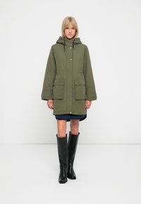 Olivengrøn parka med hætte, trykknapper og to forlommer, parret med knæhøje sorte støvler og en lagdelt blå nederdel.