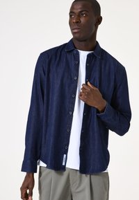 Camisa de mezclilla oscura con cuello clásico, mangas largas y un corte relajado, superpuesta sobre una camiseta blanca. Presenta un discreto detalle de etiqueta.
