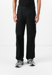 Black cotton pants with a straight-leg design, featuring a front button closure and side pockets. Worn with silver sneakers.