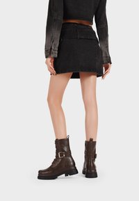 Stivali alla caviglia in pelle marrone con una finitura testurizzata, chiusura con lacci e dettaglio con fibbia laterale. Mini gonna in denim nera e giacca cropped.