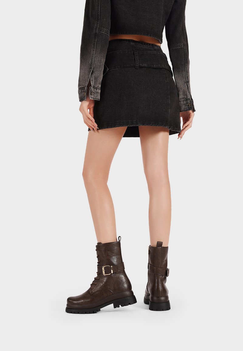 Stivali alla caviglia in pelle marrone con una finitura testurizzata, chiusura con lacci e dettaglio con fibbia laterale. Mini gonna in denim nera e giacca cropped.