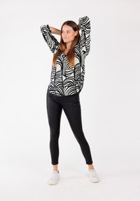 Zwart-wit gebloemde blouse met lange mouwen, gecombineerd met zwarte leggings en witte sneakers, staand tegen een witte achtergrond.