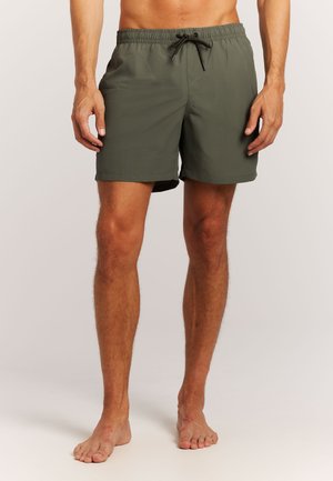 Męski model noszący oliwkowo-zielone kąpielówki z czarnym sznurkiem, stojący boso na jednolitym jasnym tle.