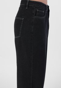 Jean noir en denim avec une coupe droite, présentant une poche latérale visible et des coutures blanches contrastées. Texture lisse avec un minimum de détails.