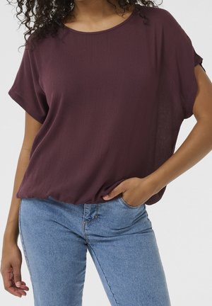 Femme portant un haut bordeaux ample à manches courtes et un jean bleu clair, une main dans la poche et des cheveux bouclés partiellement visibles.