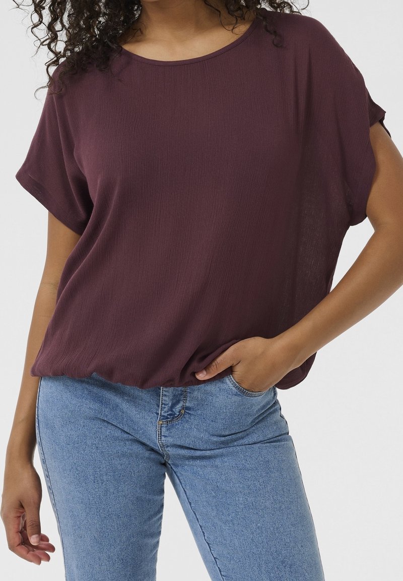 Femme portant un haut bordeaux ample à manches courtes et un jean bleu clair, une main dans la poche et des cheveux bouclés partiellement visibles.