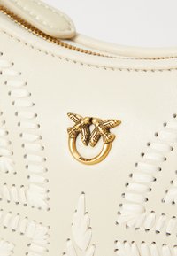 Κρέμα δερμάτινη τσάντα χειρός με περίπλοκα μοτίβα κοπής, χρυσά εξαρτήματα με δύο σχέδια πεταλούδας και κλείσιμο με φερμουάρ.