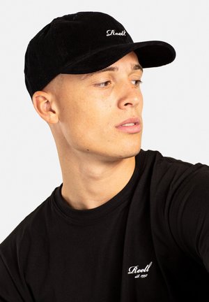 Schwarze Cord-Baseballmütze mit einem gebogenen Schirm, ausgestattet mit einem weißen gestickten Logo. Das Model trägt ein schwarzes T-Shirt mit kleinem Logo auf der Brust.