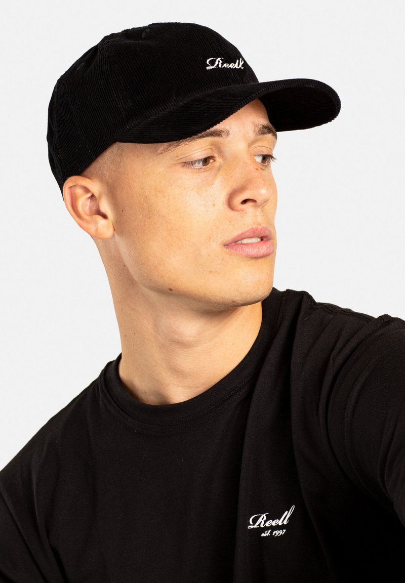Black corduroy baseball cap with a curved brim, featuring white embroidered logo. Model wears a black t-shirt with small logo on the chest.