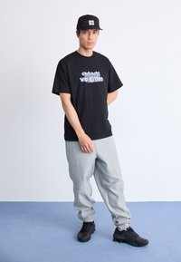 Homem com um boné preto, t-shirt gráfica preta, sweatpants cinzentas e ténis pretos, de pé sobre um chão azul contra uma parede branca.