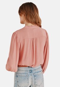 Blush-pink Bluse mit Ballonärmeln und Rüschensaum, mit lässigem Schnitt und weichem, leichtem Stoff, kombiniert mit hellen Jeans.