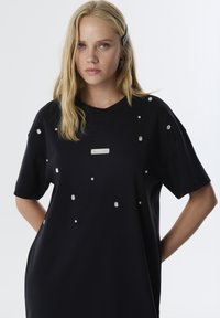 Schwarzes T-Shirt mit kurzen Ärmeln aus weichem Stoff, verziert mit verstreuten Strasssteinen und einem silbernen Etikett auf der Vorderseite.