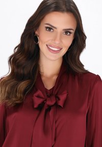 Bordowa bluzka z kokardą przy szyi. Miękka, gładka faktura i długie rękawy. Modelka ma falujące włosy i okrągłe kolczyki.