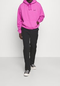 Sweat à capuche rose vif avec une poche avant et un logo Levi's noir, assorti à un pantalon noir et des baskets à motifs, présenté en vue de face.