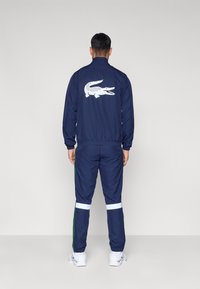 Chándal azul marino con logo blanco de cocodrilo en la espalda de la chaqueta, puños elásticos y rayas blancas en las piernas del pantalón. La tela parece suave y deportiva.