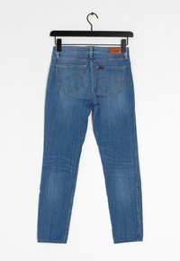 Blå denimjeans hänger från en svart klädhanger. Har raka ben, bakfickor och en brun läderlapp med "Lee"-märkning.