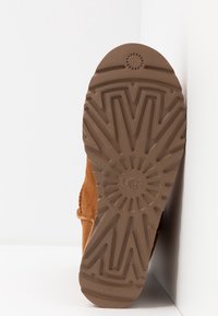 Suela de bota UGG marrón con un patrón de tracción en forma de radiación solar, que presenta el logo "UGG" y un agarre texturizado sobre un fondo blanco.