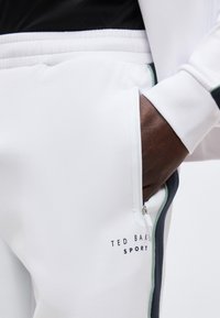 Χέρι με σκούρο δέρμα στην τσέπη με φερμουάρ άσπρου παντελονιού Ted Baker Sport με μαύρη και πράσινη πλαϊνή ρίγα.