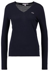 Granatowa koszula z długim rękawem i dekoltem w kształcie litery V, z małym białym haftowanym logo krokodyla po lewej stronie klatki piersiowej.