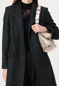 Tmavě zelený kabát z tvídu s texturovaným povrchem, jednorzadý střih a klopy s výstřihem do V. Drží kovovou zlatou tašku s plisováním.