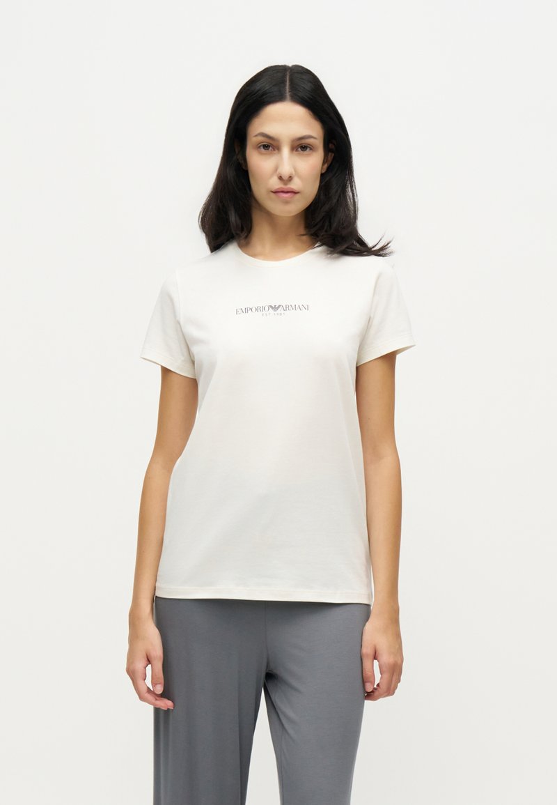 Γυναίκα με σκούρα μαλλιά που φοράει λευκό t-shirt Emporio Armani και γκρι παντελόνι, στέκεται μπροστά σε απλό λευκό φόντο.