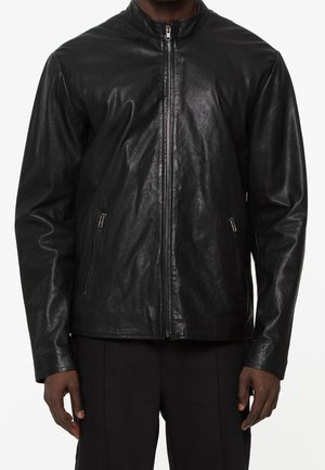 Homme portant une veste en cuir noire avec une fermeture éclair frontale et des poches latérales zippées, associée à un pantalon noir.