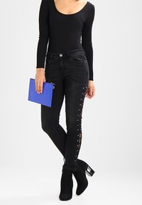 Jeans ajustados negros con detalles de encaje en los laterales, combinados con una blusa de manga larga ceñida y un bolso de mano azul. Botines negros de tacón completan el conjunto.