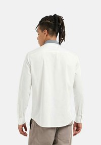 Camicia bianca a maniche lunghe con colletto blu a contrasto, design pulito e orlo leggermente curvato, realizzata in tessuto leggero.